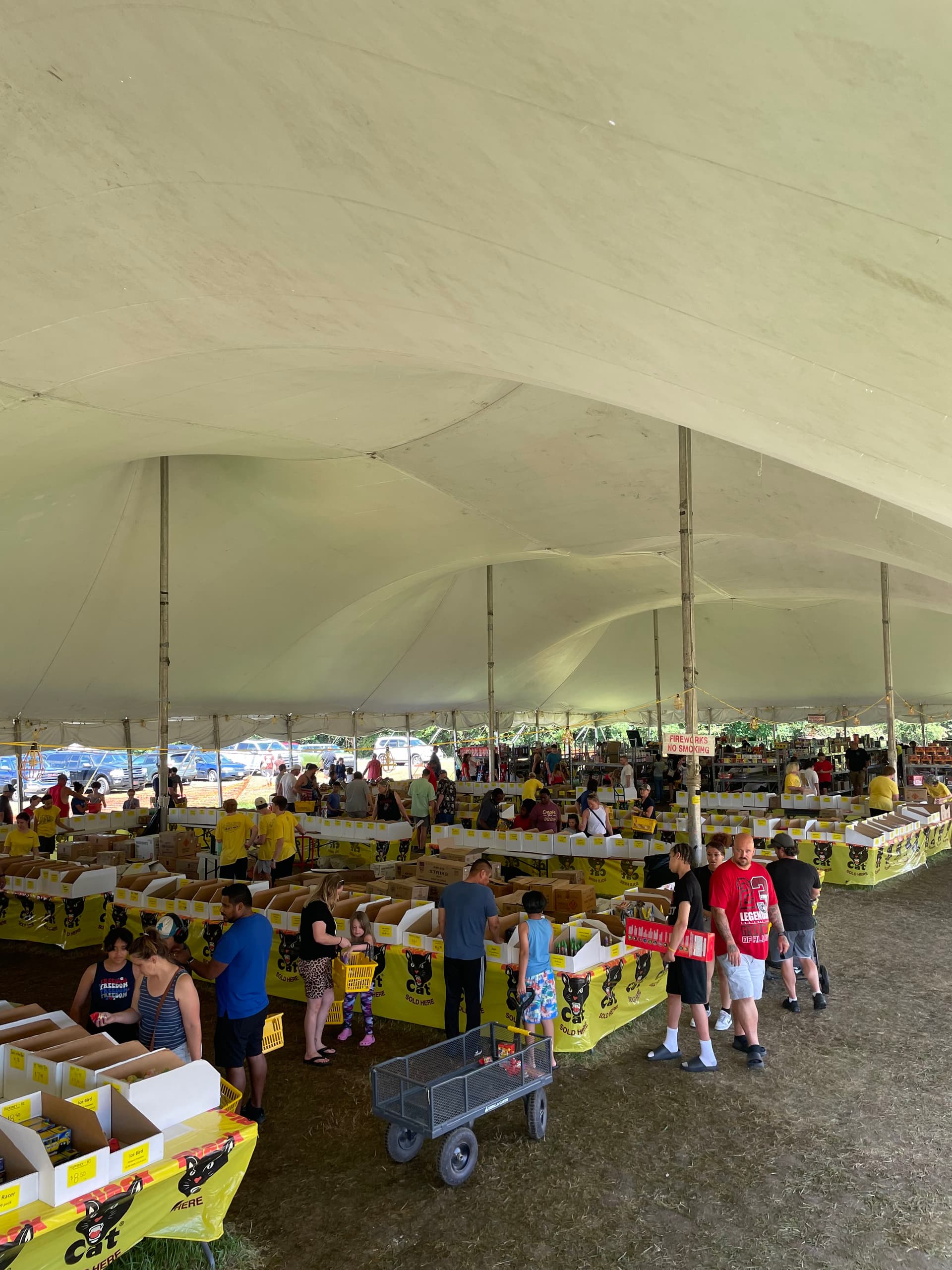 Inside Crazy Joe's 200-foot Kansas City fireworks tent — staff in red Crazy Joe's shirts working the long aisle of yellow Black Cat fireworks tables piled high with artillery shells, 500g cakes, and assortment packs