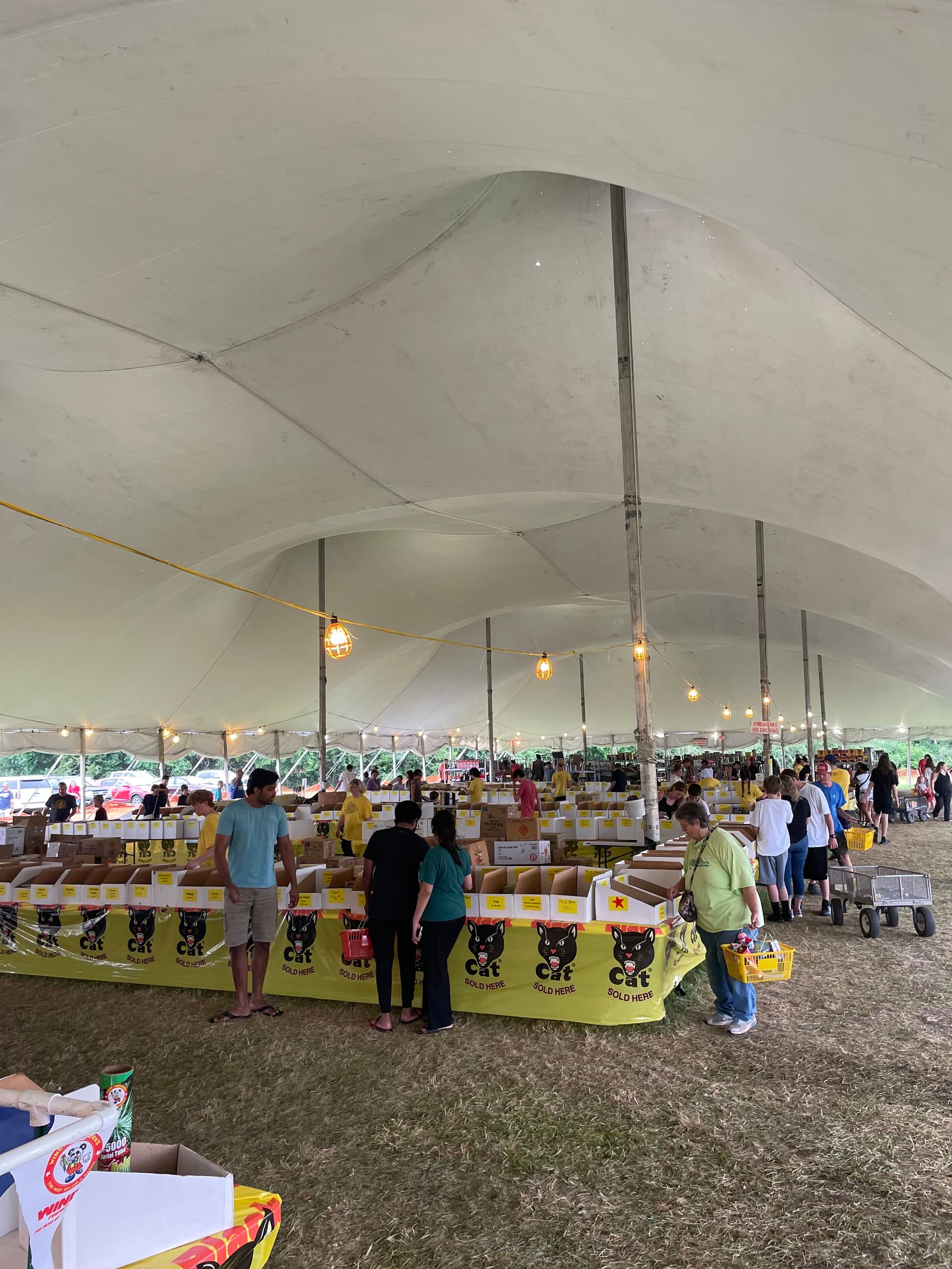 Crazy Joe's Fireworks shoppers, including a child, browsing the 200-foot aisle of yellow Black Cat fireworks tables under string lights inside the white event tent at 171st and Holmes in Kansas City