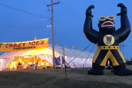 Crazy Joe's Fireworks stand at dusk — the giant inflatable Joerilla gorilla mascot stands beside the lit-up white event tent, the yellow CRAZY JOE'S FIREWORKS banner over the entrance, customers visible inside the tent