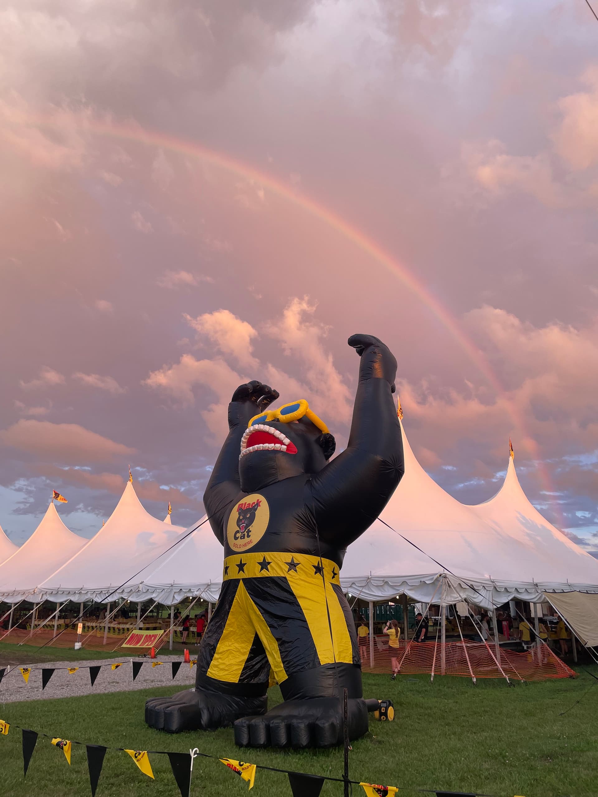 Iconic dusk view of Crazy Joe's Fireworks — the giant inflatable Joerilla mascot in profile with a full rainbow arching across the sunset sky above the white event tents at 171st & Holmes in Kansas City